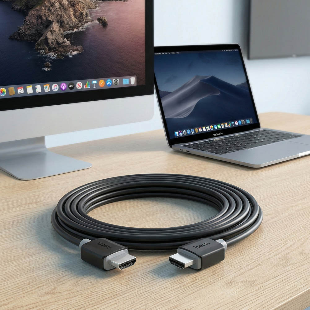 Cable HDMI HOCO negro de 1 metro con conectores HDMI en ambos extremos, junto a un iMac y un MacBook sobre un escritorio.