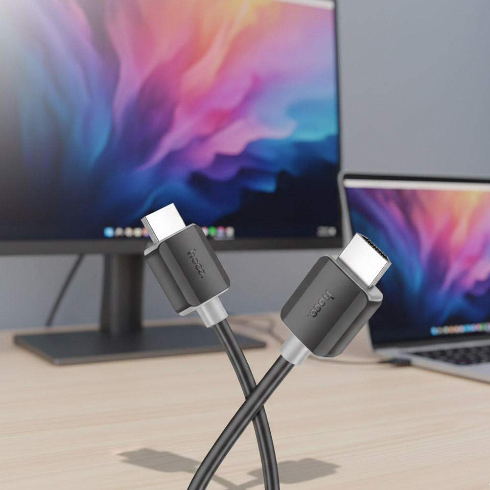 Cable HDMI HOCO negro con conectores metálicos, presentado junto a un monitor y un portátil para transmisión de video en alta definición.