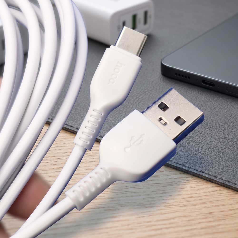 Un cable Hoco X20 blanco de 3 metros con conector USB-A a USB-C aparece junto a un cargador y un teléfono sobre una mesa.