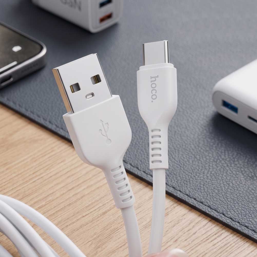 Un cable USB a USB-C blanco Hoco X20 muestra conectores reforzados junto a una base de carga sobre una mesa de madera.