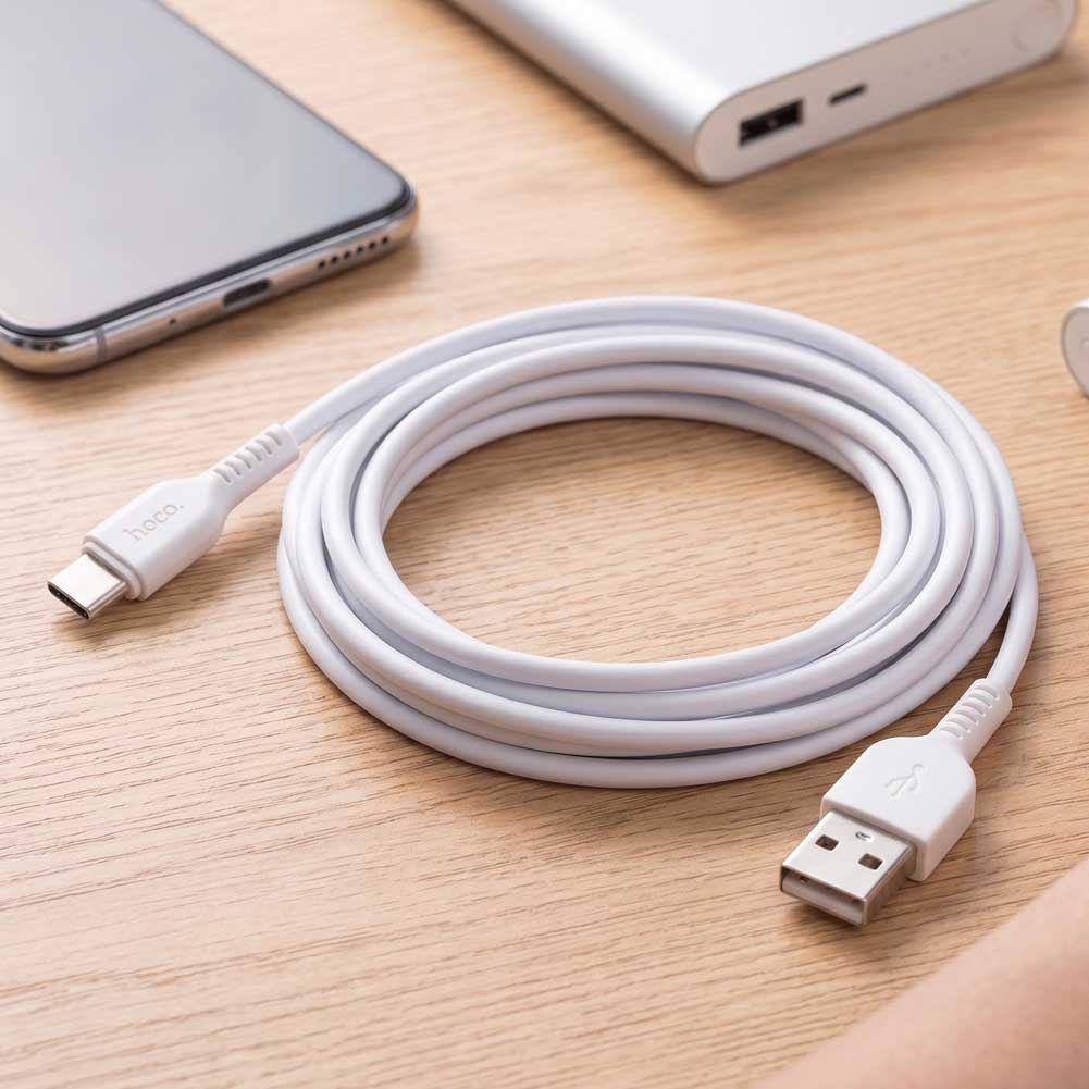 Cable Hoco X20 blanco de 3 metros con conector USB-A y USB-C sobre una mesa de madera, junto a un teléfono y una batería externa.