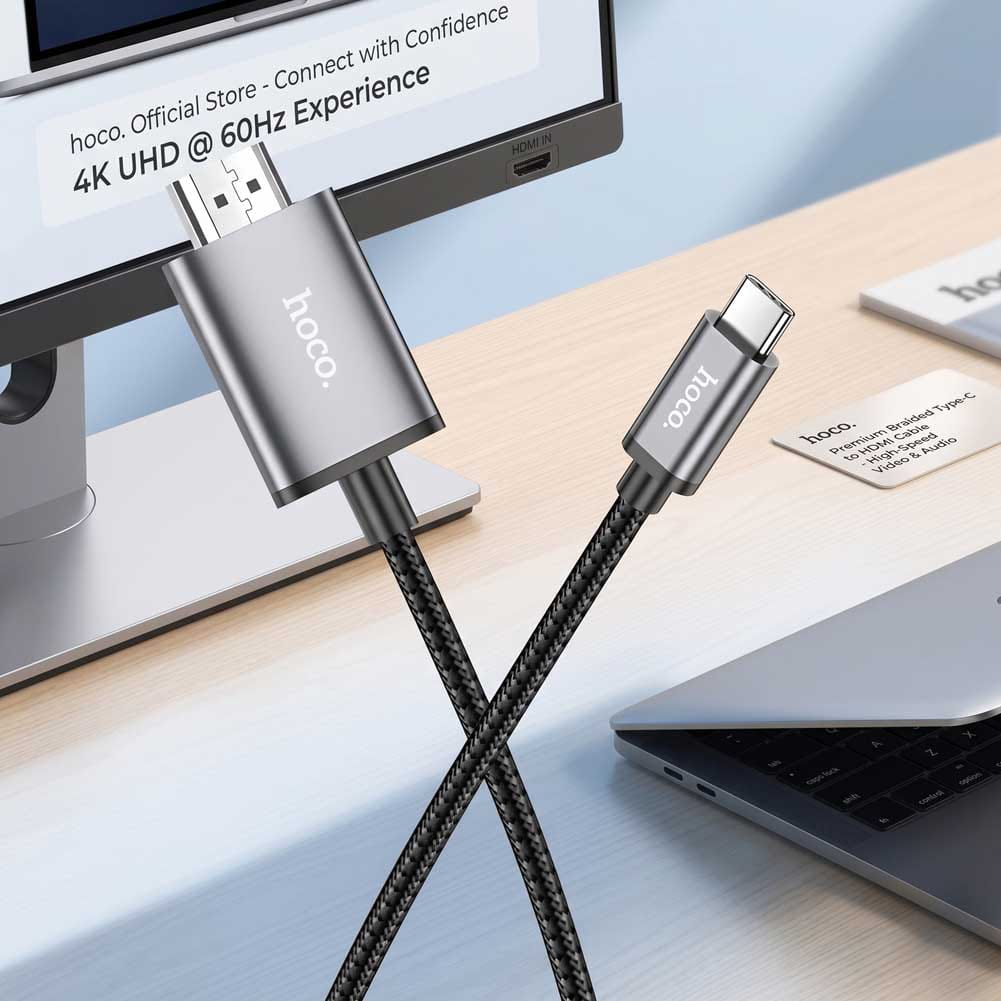 Un cable Hoco USB-C a HDMI de color negro conecta una laptop con un monitor, con texto visible sobre 4K UHD y acabado trenzado.