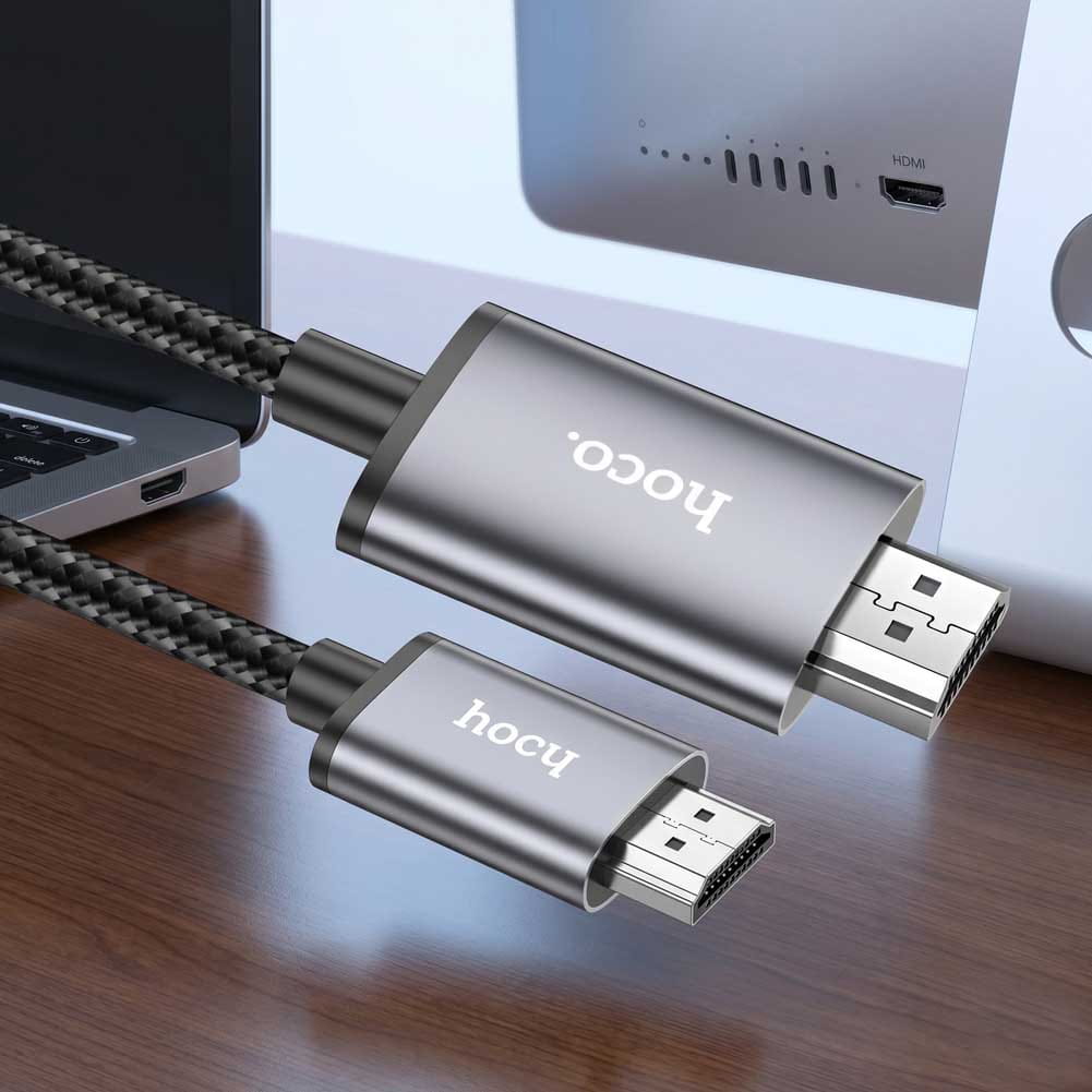 Un cable Hoco negro trenzado conecta USB-C y HDMI junto a un portátil y una pantalla, con conectores metálicos y acabado resistente 4K.
