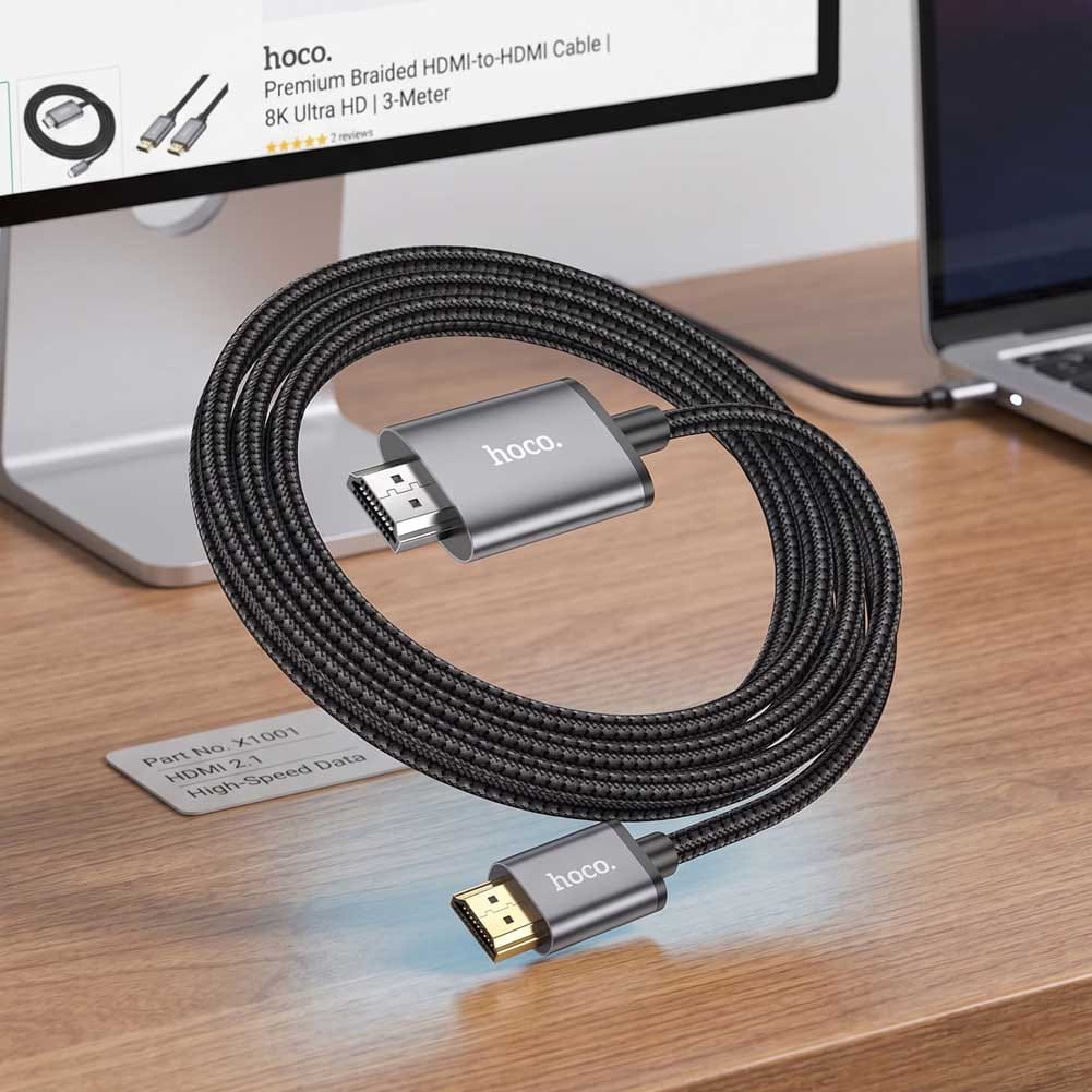 Un cable Hoco USB-C a HDMI negro y trenzado conecta un portátil con un monitor, con conectores metálicos y acabado de 2 metros.