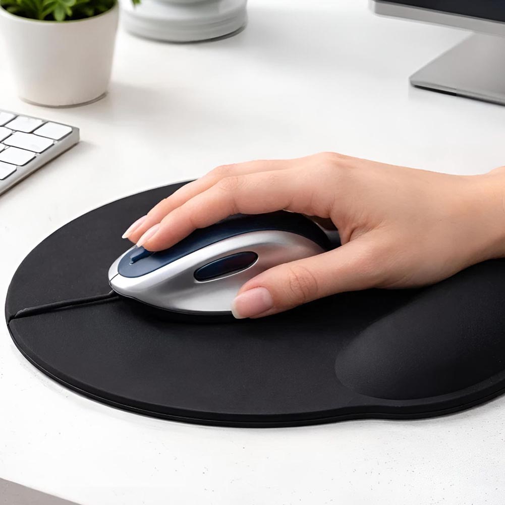 Una mano usa un mouse sobre un mouse pad negro ergonómico con apoyo para la muñeca y base antideslizante, en un escritorio blanco junto a un teclado.
