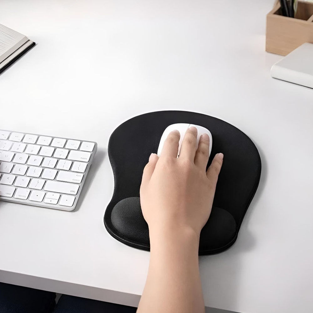 Una mano descansa sobre un mouse pad ergonómico negro con apoyo de gel en una mesa blanca, junto a un teclado compacto y accesorios de escritorio.