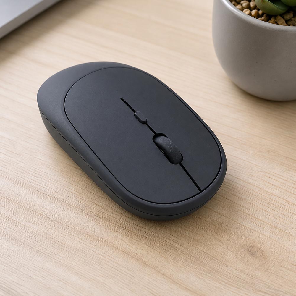 Un mouse inalámbrico negro y slim descansa sobre una mesa de madera clara junto a una maceta decorativa, con diseño recargable y 4 botones.