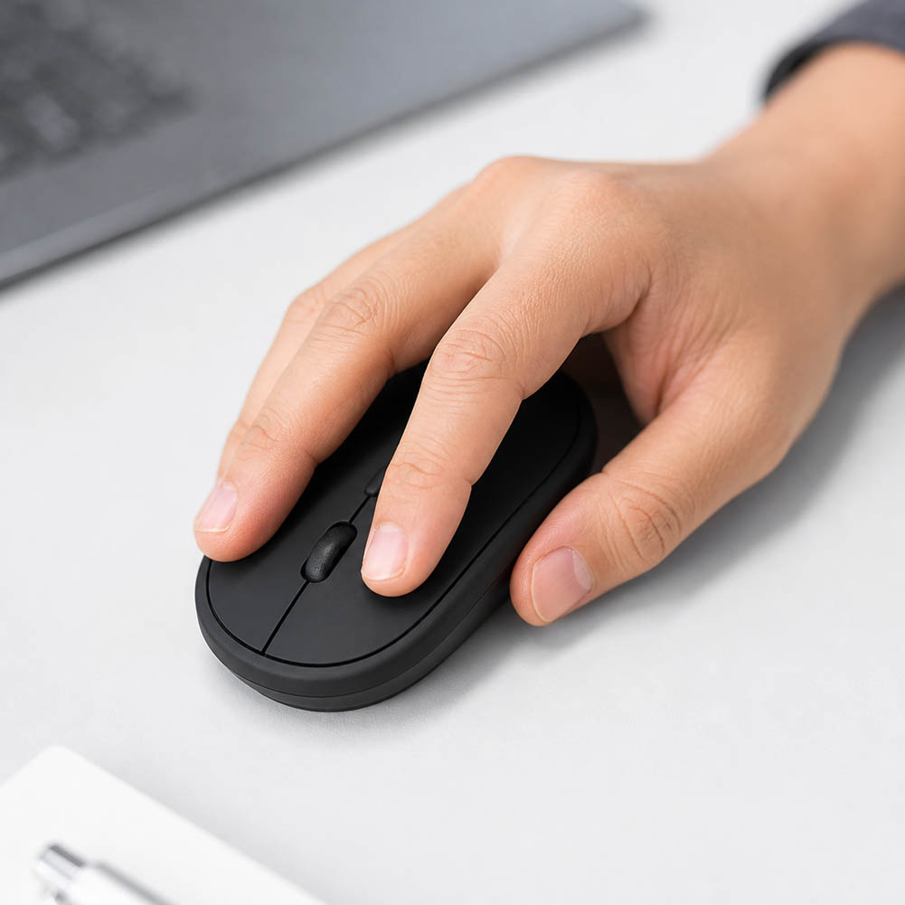 Una mano descansa sobre un mouse inalámbrico recargable negro y compacto, junto a un portátil, en una superficie blanca de escritorio.