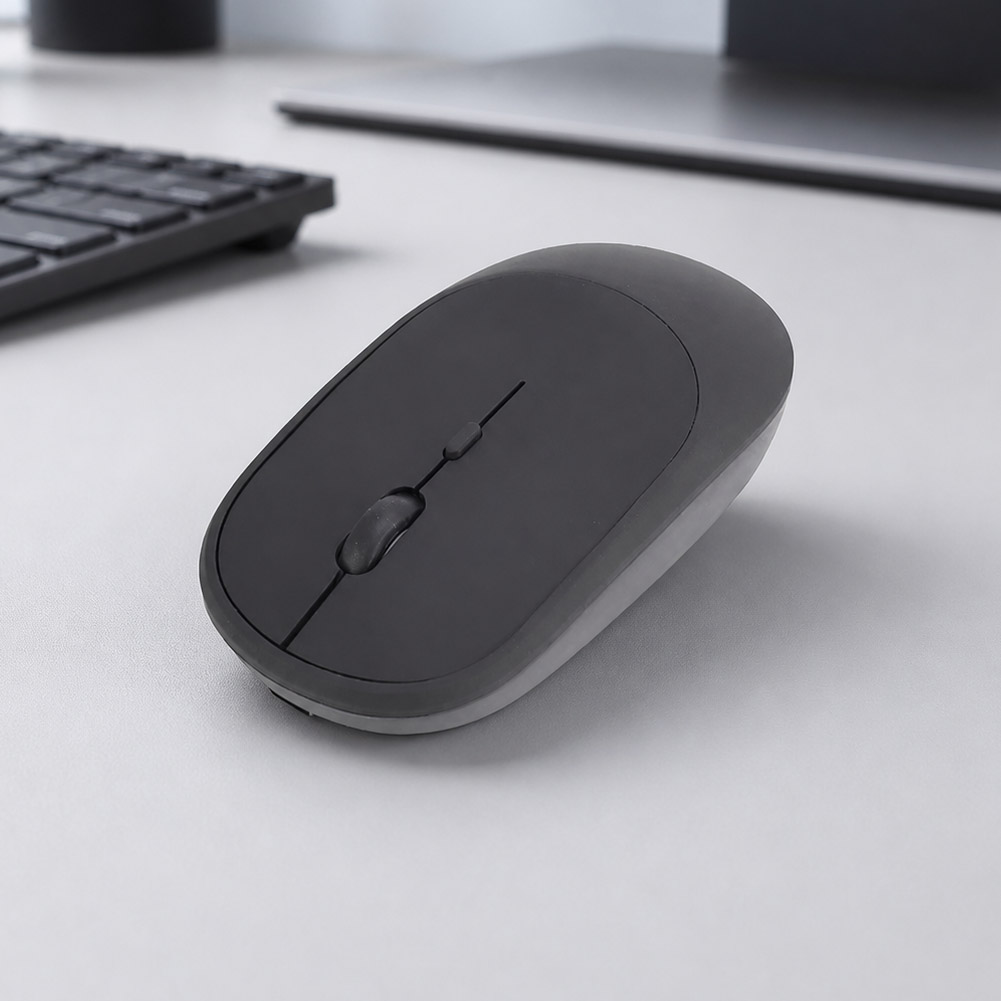 Un mouse inalámbrico recargable slim negro descansa sobre un escritorio claro junto a un teclado, con diseño compacto y botones visibles.