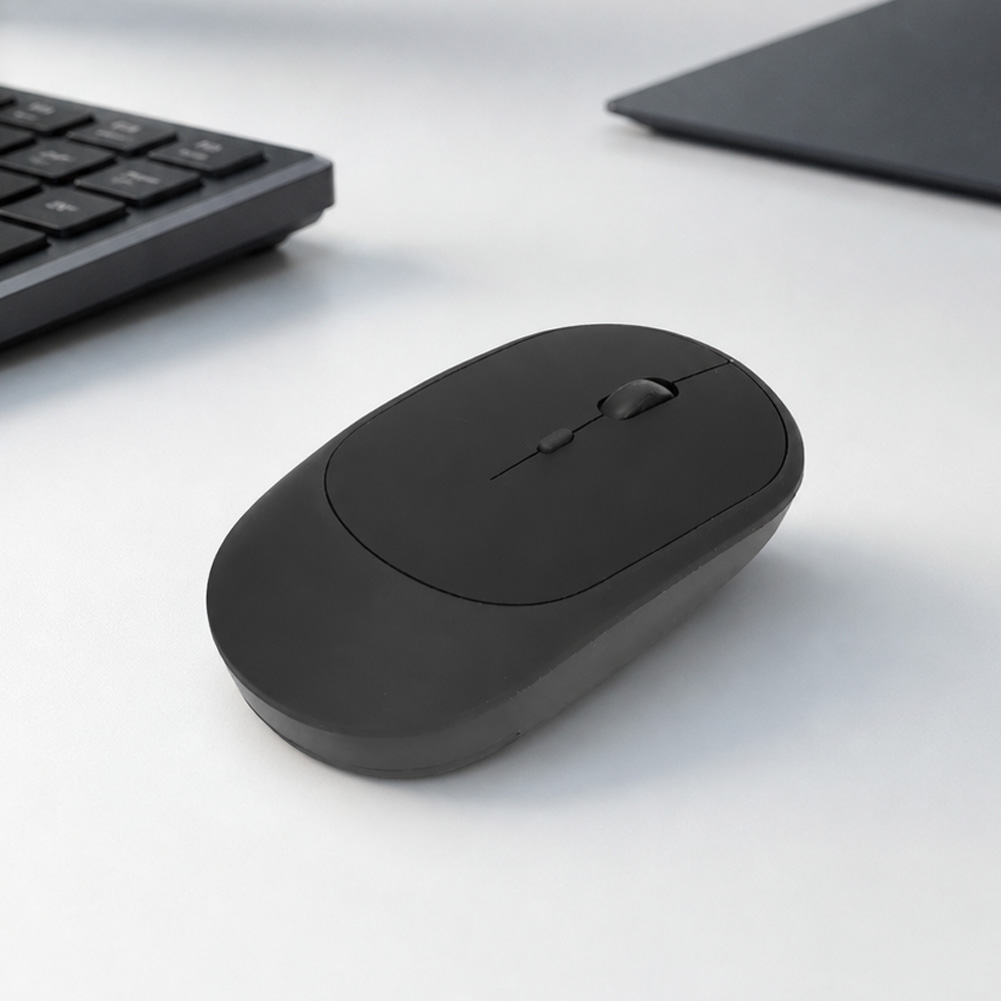 Un mouse inalámbrico negro y recargable descansa sobre un escritorio blanco, junto a un teclado y un portátil en tono sobrio.