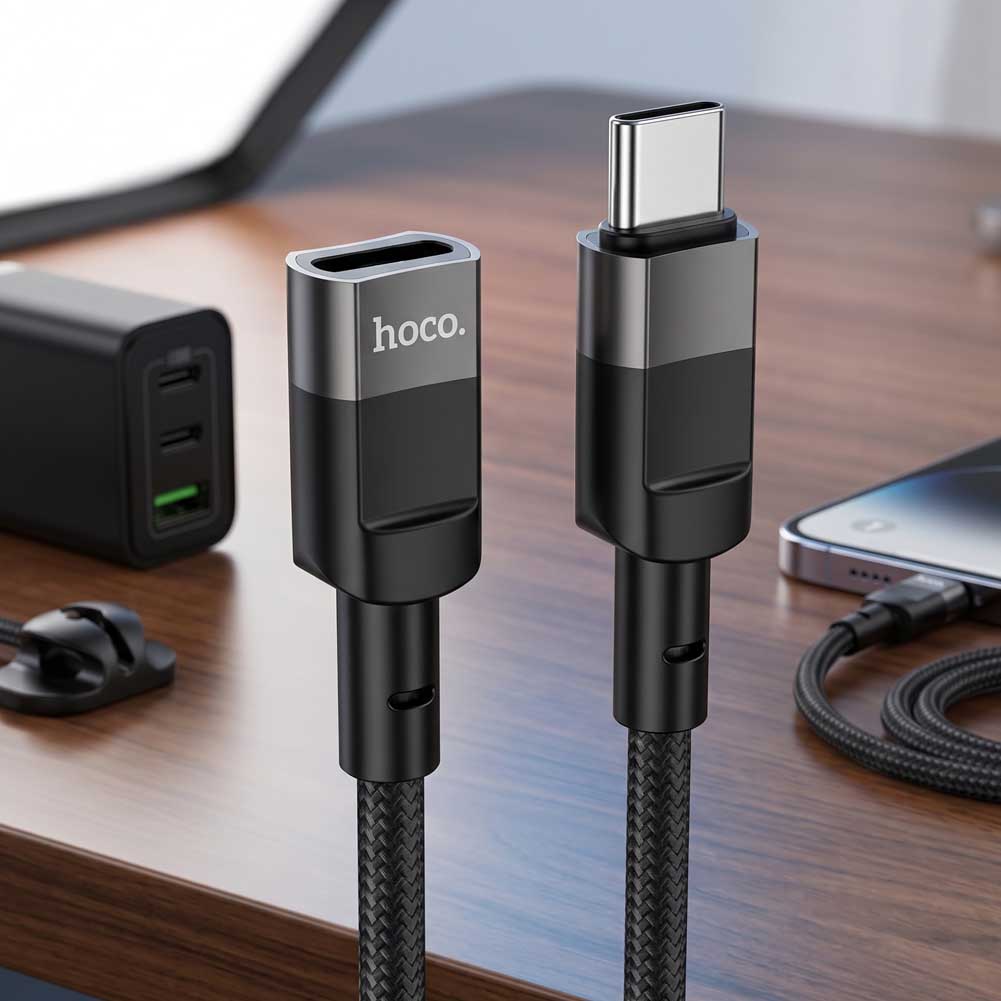 En un escritorio, el cable Hoco U107 USB-C negro con conectores de aluminio extiende carga y transferencia de datos entre dispositivos de forma práctica.
