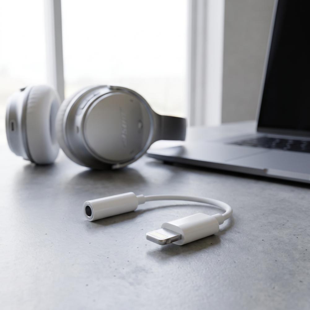 Adaptador blanco de Lightning a jack de 3,5 mm junto a unos auriculares y un portátil sobre una mesa.